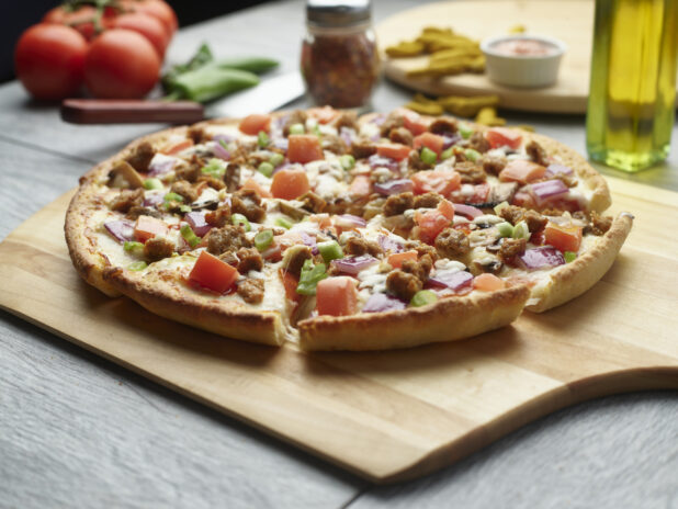 Sliced pizza with sausage, tomatoes, mushrooms and red onion topped with fresh green onion on a wooden cutting board surrounded by ingredients in the background