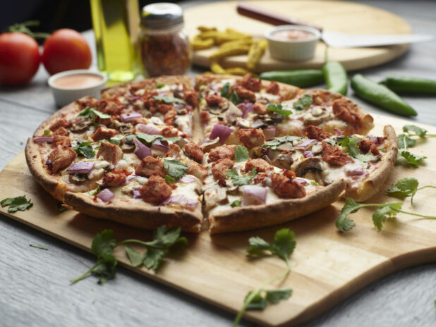 Sliced tandoori chicken pizza with mushrooms, red onion and chicken topped with cilantro on a wooden peel surrounded by ingredients