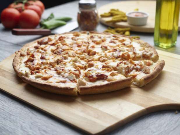 Sliced barbecue chicken pizza on a wooden cutting board surrounded by ingredients in the background