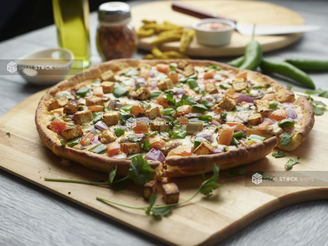 Sliced paneer tikka masala pizza with mushrooms, red onion, peppers and tomatoes topped with fresh cilantro on a wooden cutting board surrounded by ingredients in the background