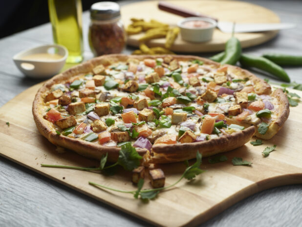 Sliced paneer tikka masala pizza with mushrooms, red onion, peppers and tomatoes topped with fresh cilantro on a wooden cutting board surrounded by ingredients in the background
