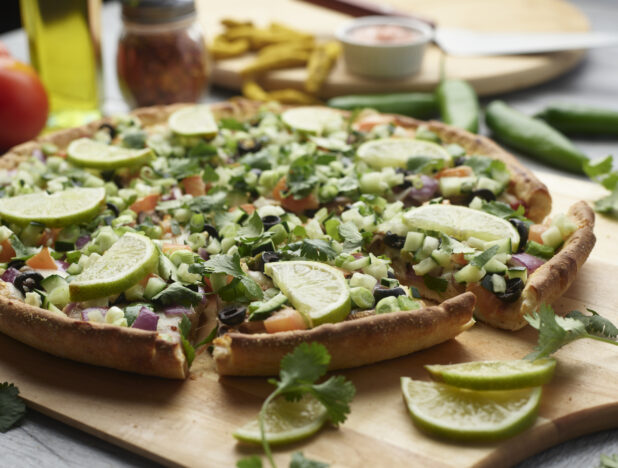 Sliced vegetarian pizza topped with fresh slices of lime on a wooden cutting board surrounded by ingredients in the background