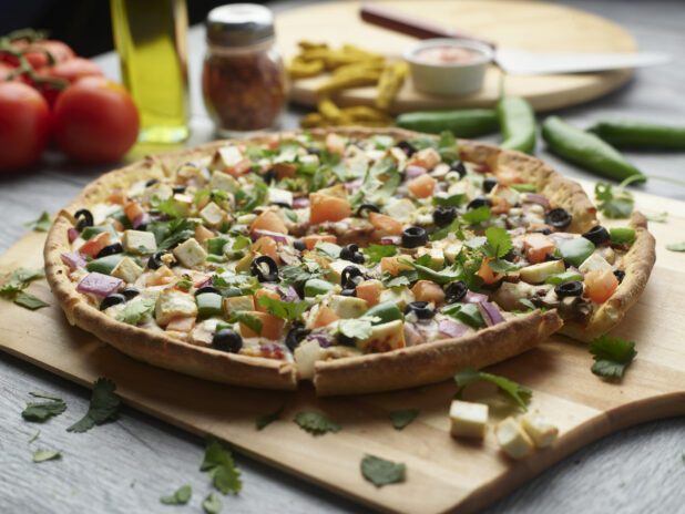 Sliced paneer tikka masala pizza on a wooden cutting board surrounded by ingredients in the background