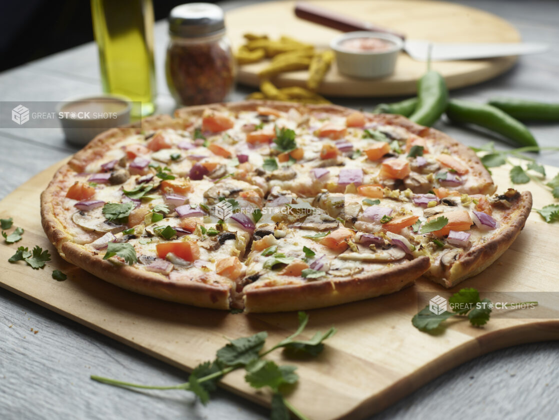 Sliced tandoori veggie pizza with mushrooms, red onion and tomatoes topped with fresh cilantro on a wooden cutting board surrounded by ingredients in the background