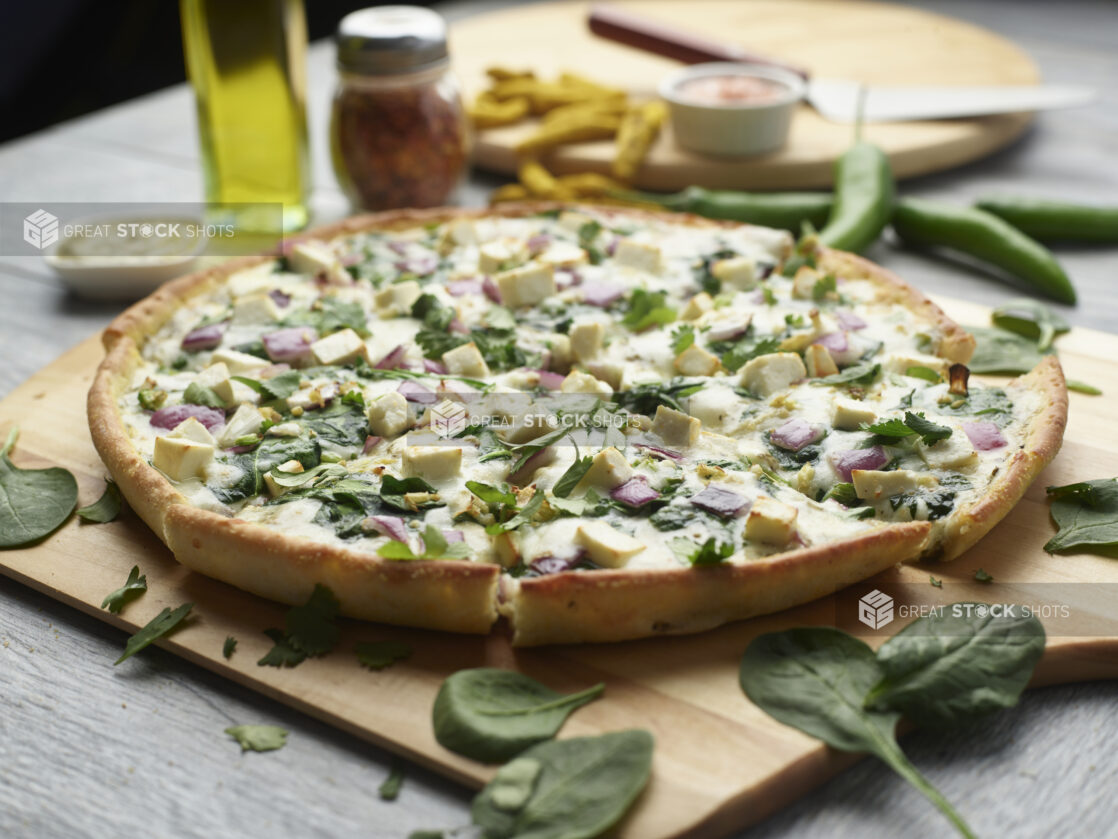 Sliced palak paneer pizza with pesto, spinach and red onion topped with fresh cilantro on a wooden cutting board surrounded by ingredients in the background