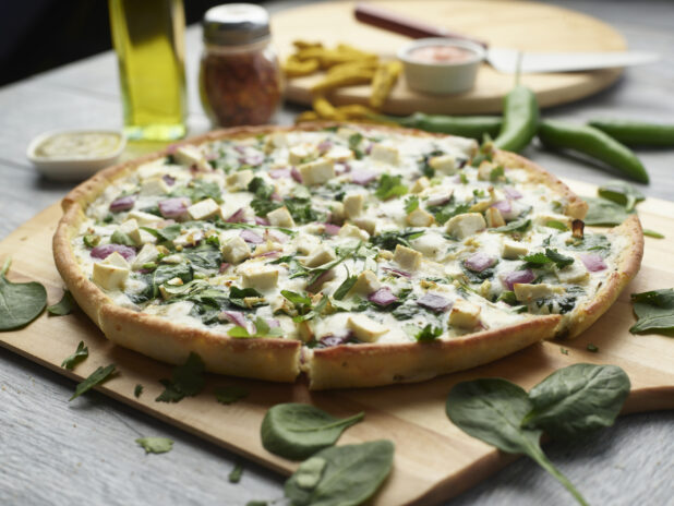 Sliced palak paneer pizza with pesto, spinach and red onion topped with fresh cilantro on a wooden cutting board surrounded by ingredients in the background