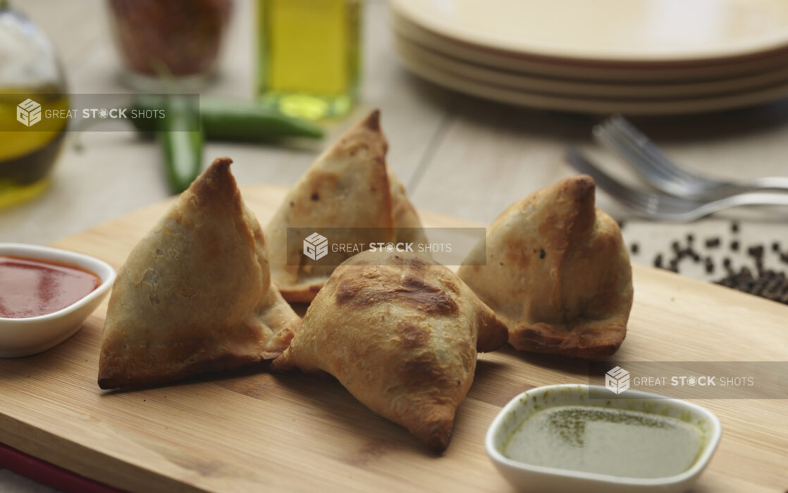 Four stuffed samosas with red and green dipping sauces on a wooden cutting board