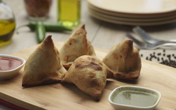 Four stuffed samosas with red and green dipping sauces on a wooden cutting board