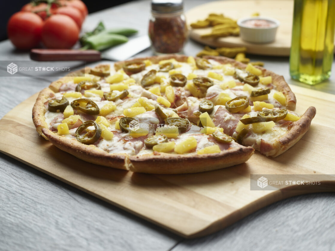 Sliced spicy Hawaiian pizza with ham, jalapeño and pineapple on a wooden cutting board surrounded by ingredients in the background