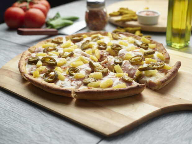 Sliced spicy Hawaiian pizza with ham, jalapeño and pineapple on a wooden cutting board surrounded by ingredients in the background