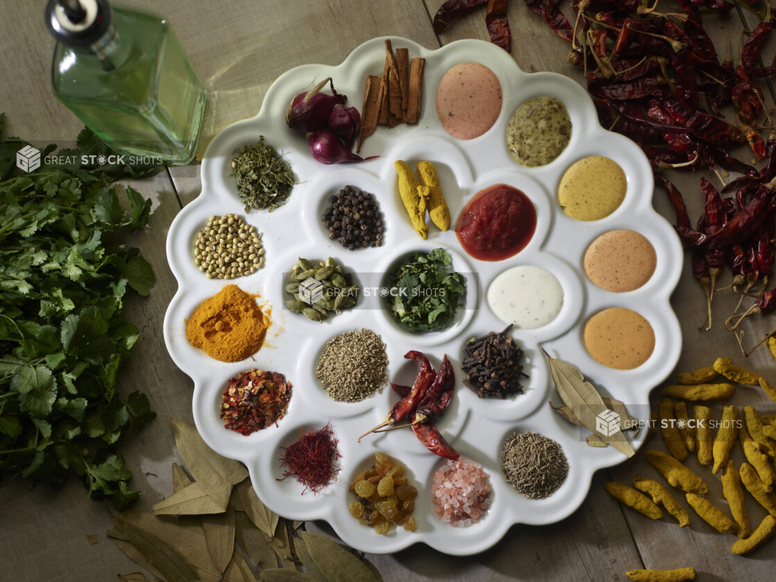 Overhead tray of Indian spices surrounded by fresh and dried ingredients on a wooden background