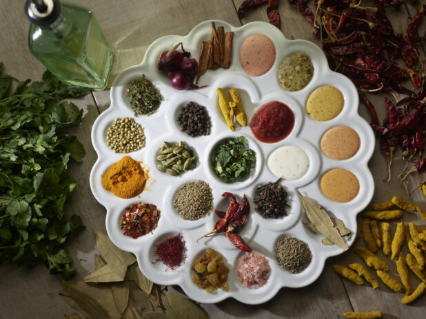 Overhead tray of Indian spices surrounded by fresh and dried ingredients on a wooden background