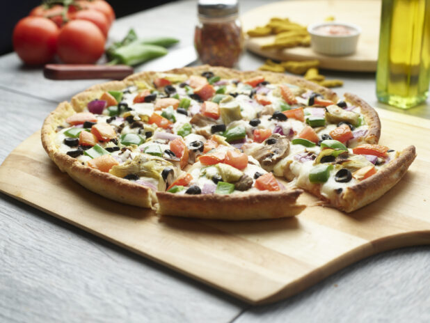 Sliced veggie pizza on a wooden cutting board surrounded by ingredients in the background