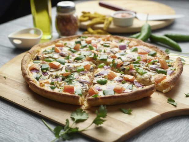 Sliced tikka masala pizza with mushrooms, red onion, peppers and tomatoes topped with fresh cilantro on a wooden cutting board surrounded by ingredients in the background