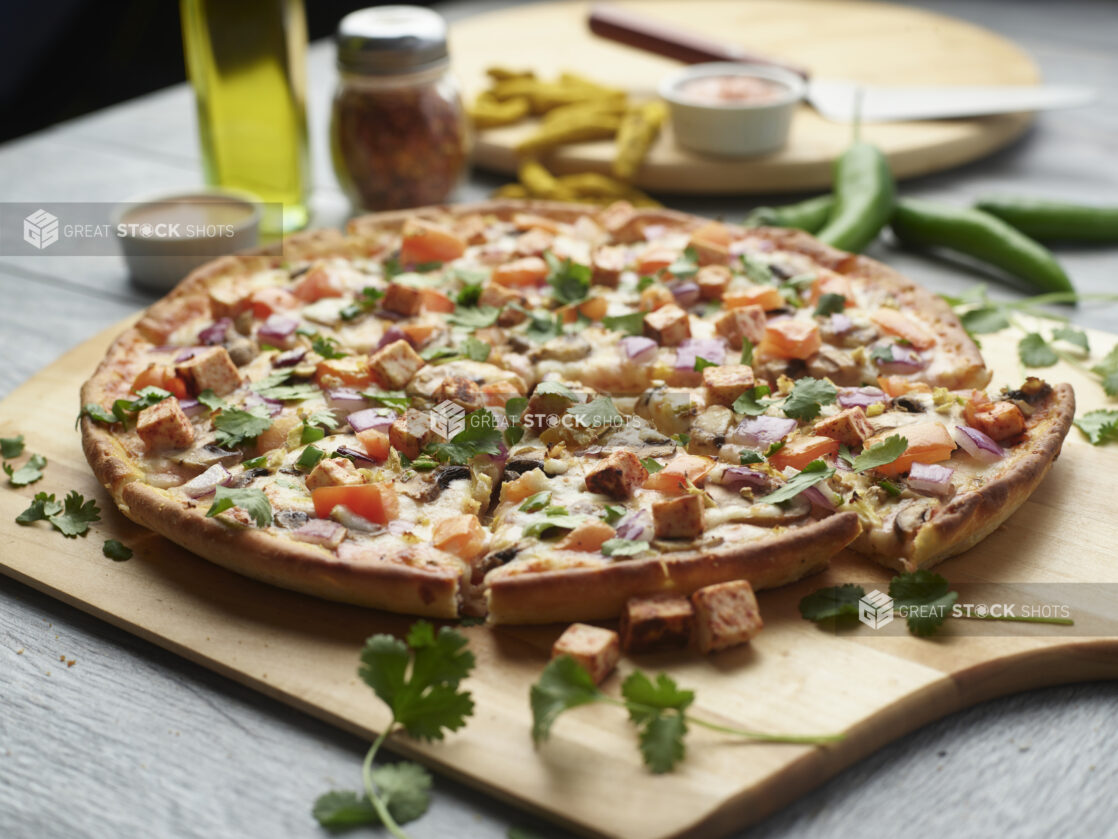 Sliced tandoori paneer pizza with mushrooms, red onion and tomatoes topped with fresh cilantro on a wooden cutting board surrounded by ingredients in the background