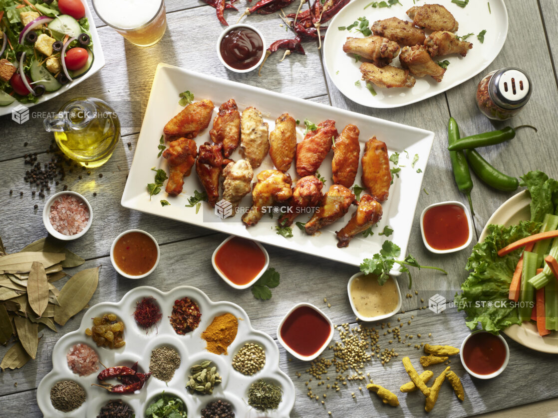 Overhead platter of various flavored chicken wings surrounded with different dipping sauces and dried herbs and spices on a wooden background
