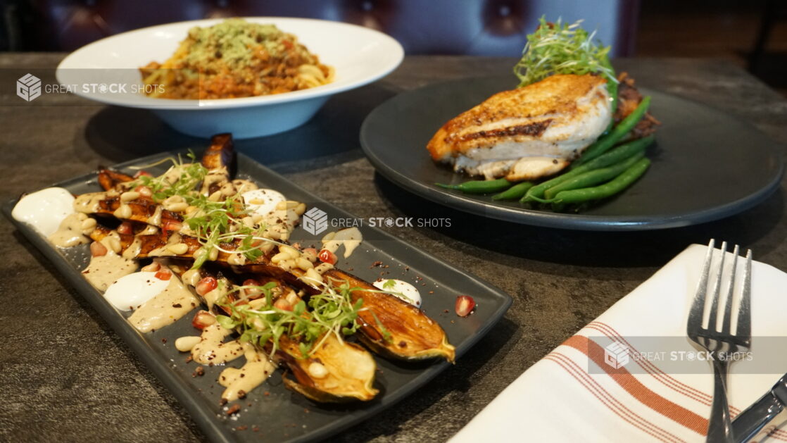 Grilled eggplant halves drizzled in sauce with pomegranate seeds with two entrees of roasted chicken and beans and spaghetti bolognese on a black slate table in a restaurant