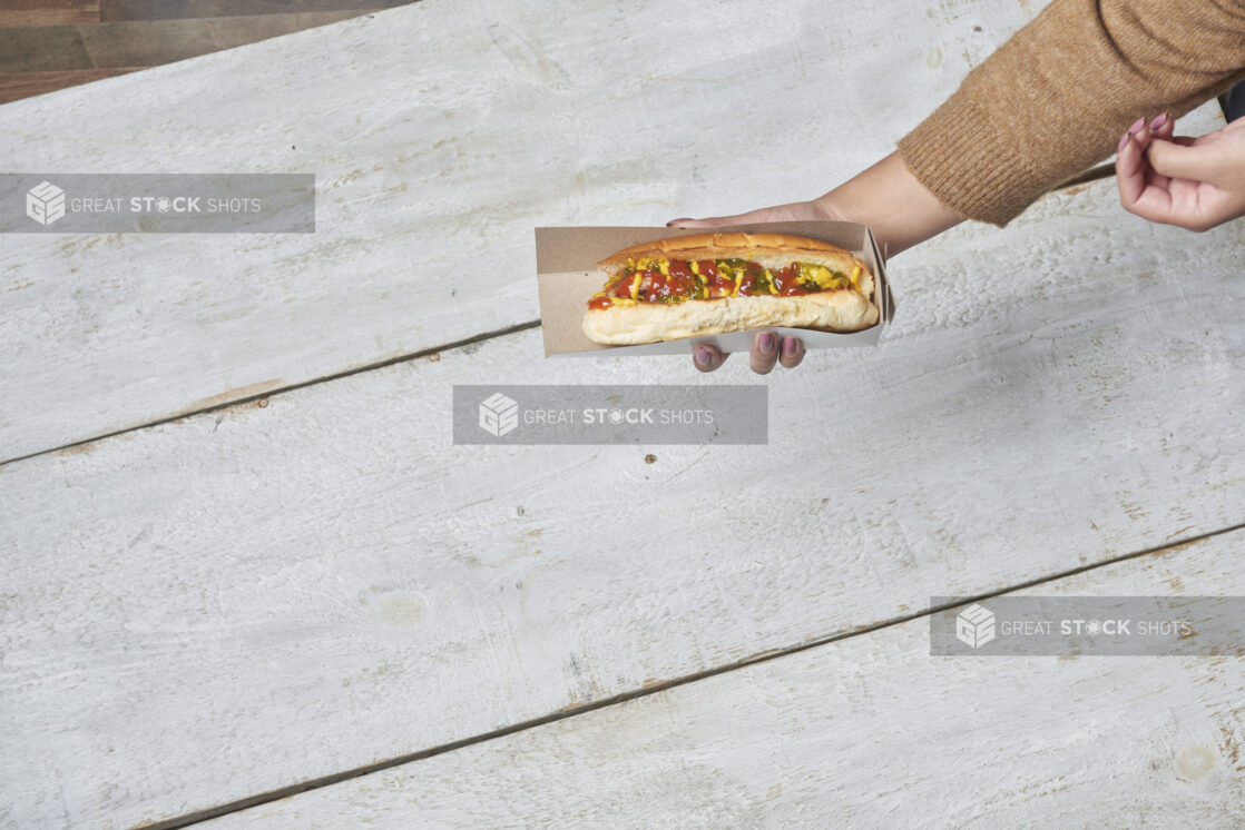 Top down of a women holding a hot dog in a cardboard sleeve with ketchup, mustard and relish over a wood table