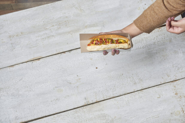 Top down of a women holding a hot dog in a cardboard sleeve with ketchup, mustard and relish over a wood table