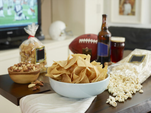 Large white bowl of tortilla chips with popcorn, peanuts, beer and a football on a wooden table in a living room with football on TV in the background