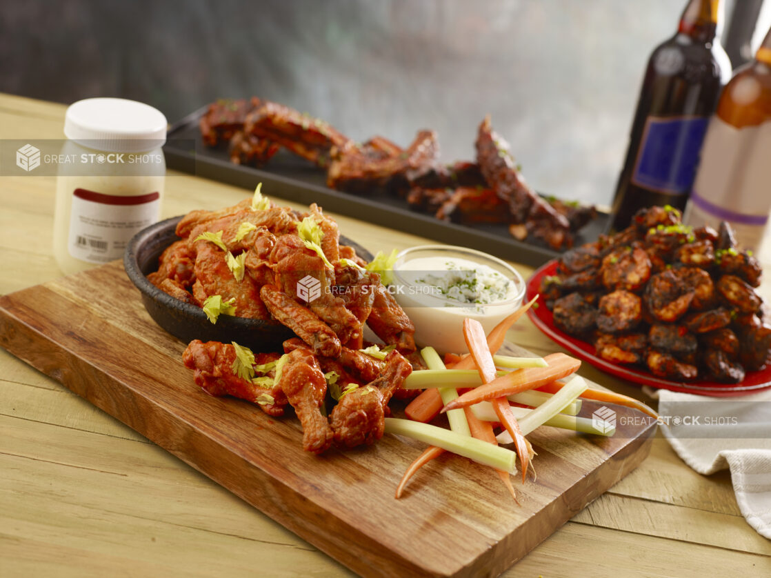 Sauced chicken wings with celery, carrots and blue cheese dip with blackened shrimp and ribs in the background on a wood table