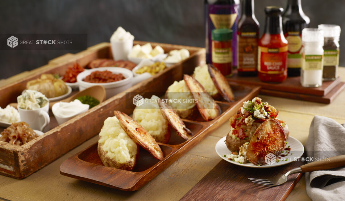 Loaded baked potato with bacon, feta cheese, sauce and chives with a baked potato parmesan chip on a board with a row of potatoes, toppings and condiments in behind