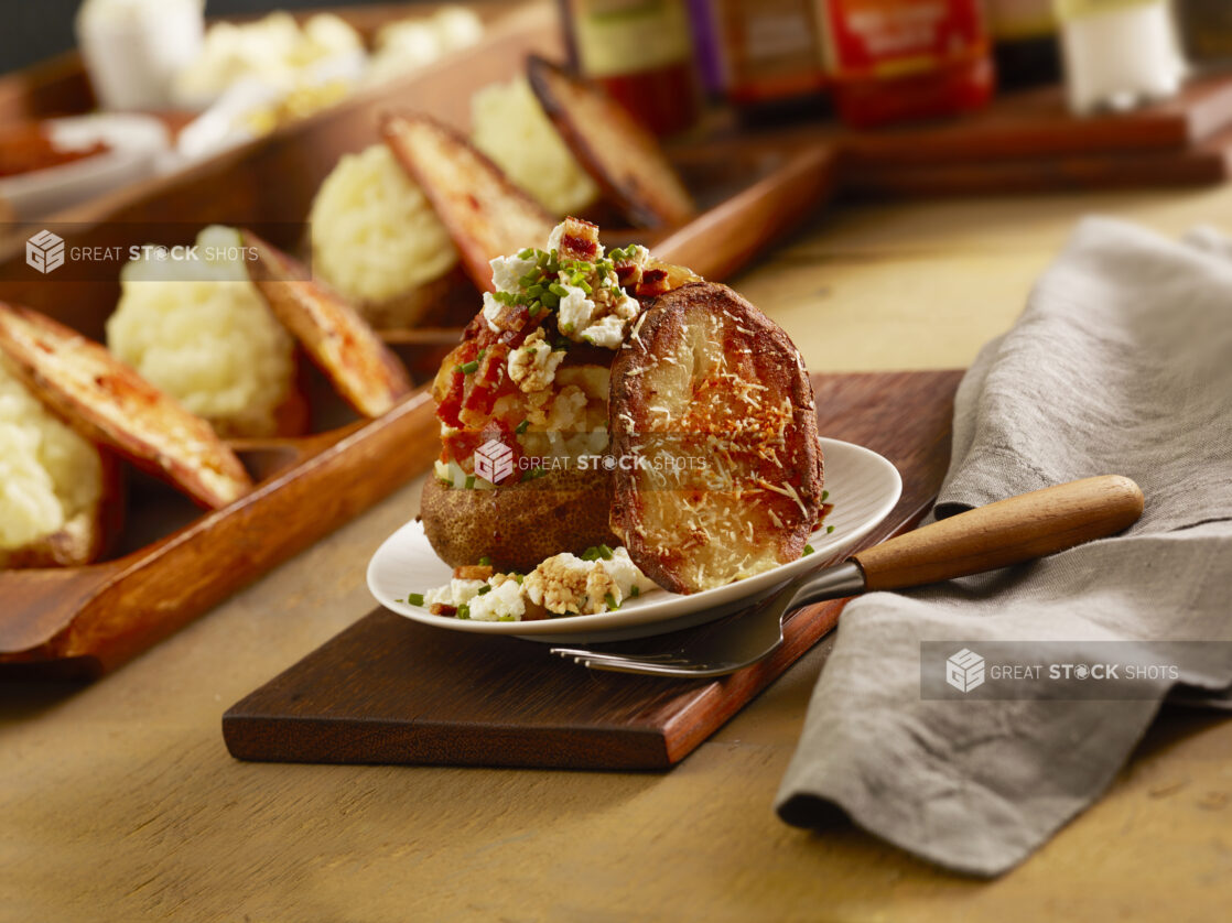 Loaded baked potato with bacon, feta cheese, sauce and chives with a baked potato parmesan chip on a board with a row of potatoes in behind