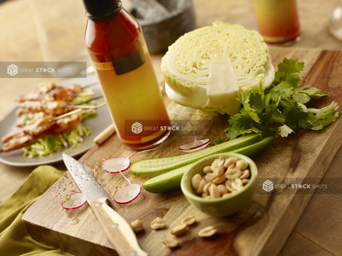 Bottle of Thai chili sauce on a wood board with cabbage, halved cucumber, parsley and a bowl of peanuts with a plate of chicken satay in the background