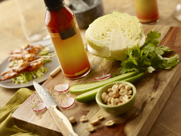 Bottle of Thai chili sauce on a wood board with cabbage, halved cucumber, parsley and a bowl of peanuts with a plate of chicken satay in the background