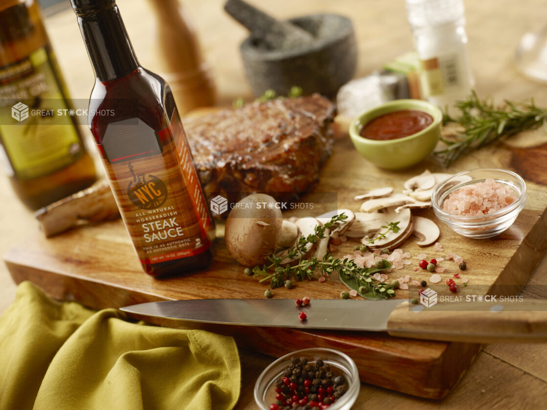 Tomahawk steak with steak sauce, fresh herbs, fresh salt and pepper, fresh mushrooms and a carving knife on a wooden board