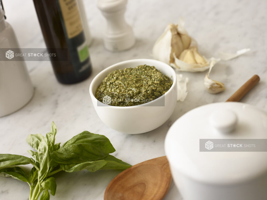 White bowl of pesto with fresh basil and garlic in the background on white marble