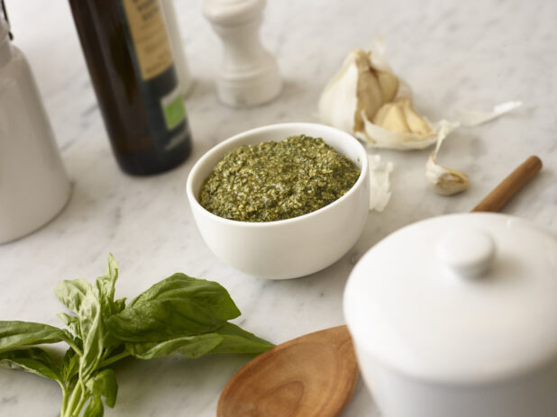 White bowl of pesto with fresh basil and garlic in the background on white marble