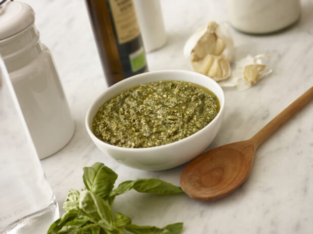 White bowl of pesto with fresh basil and garlic in the background on white marble