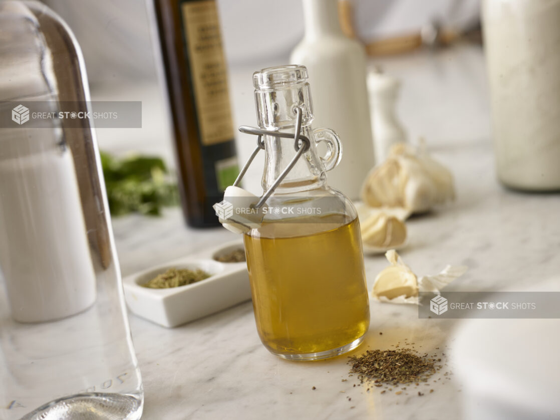 Opened glass bottle of olive oil infused with garlic, herbs and spices on a white marble countertop surrounded by ingredients