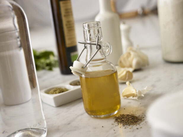 Opened glass bottle of olive oil infused with garlic, herbs and spices on a white marble countertop surrounded by ingredients