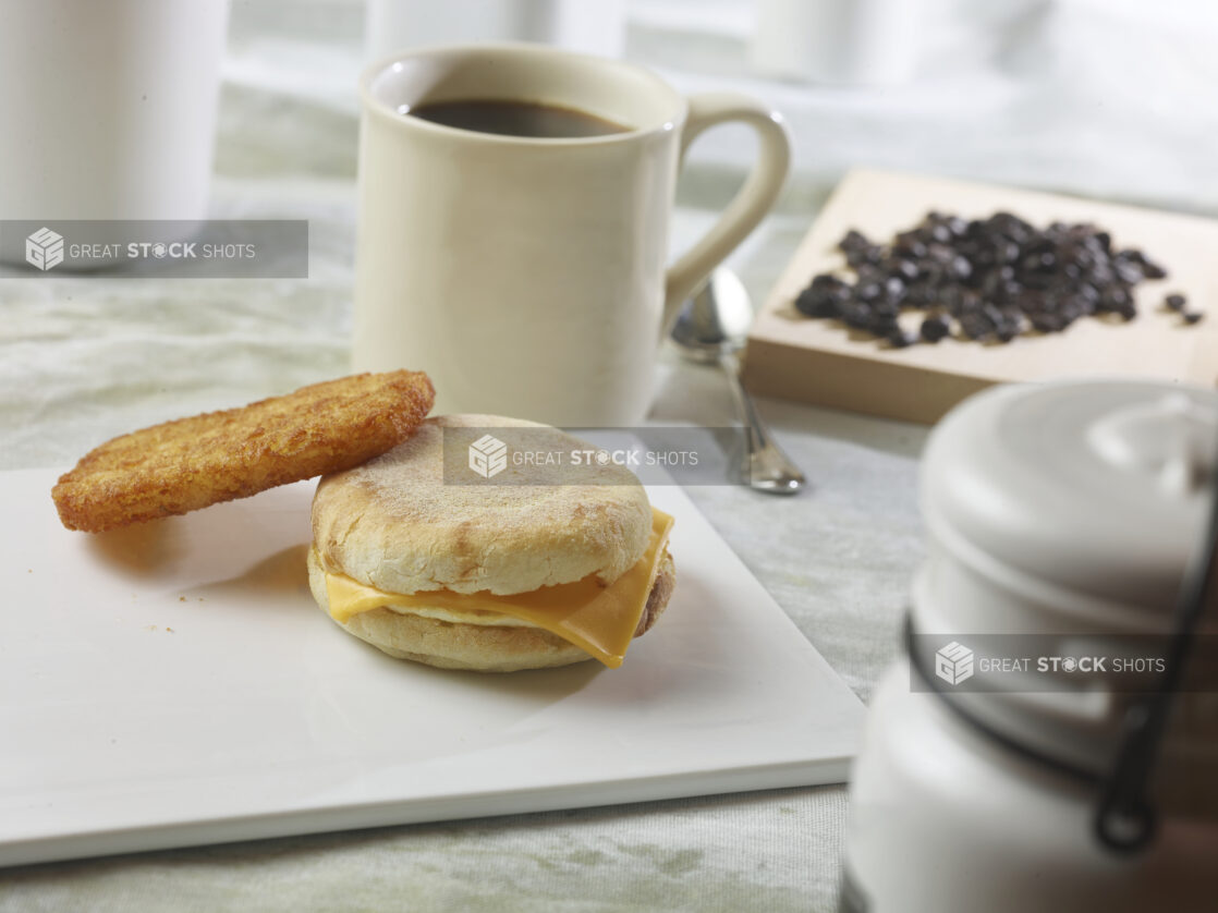 Egg and cheese breakfast sandwich with a hash brown on a white board with a cup of coffee with coffee beans in the background