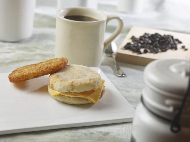 Egg and cheese breakfast sandwich with a hash brown on a white board with a cup of coffee with coffee beans in the background