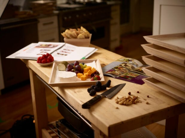 Preparing a wooden catering tray with cheese and fruit in a kitchen with instructions on a wooden table