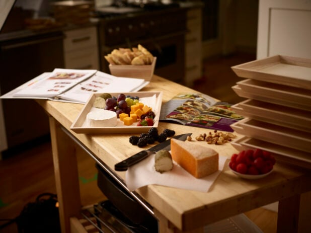 Preparing a wooden catering tray with cheese and fruit in a kitchen with instructions on a wooden table