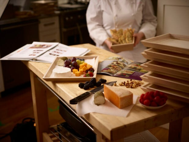 Chef holding a wooden basket of bread while preparing a wooden catering tray with cheese and fruit in a kitchen