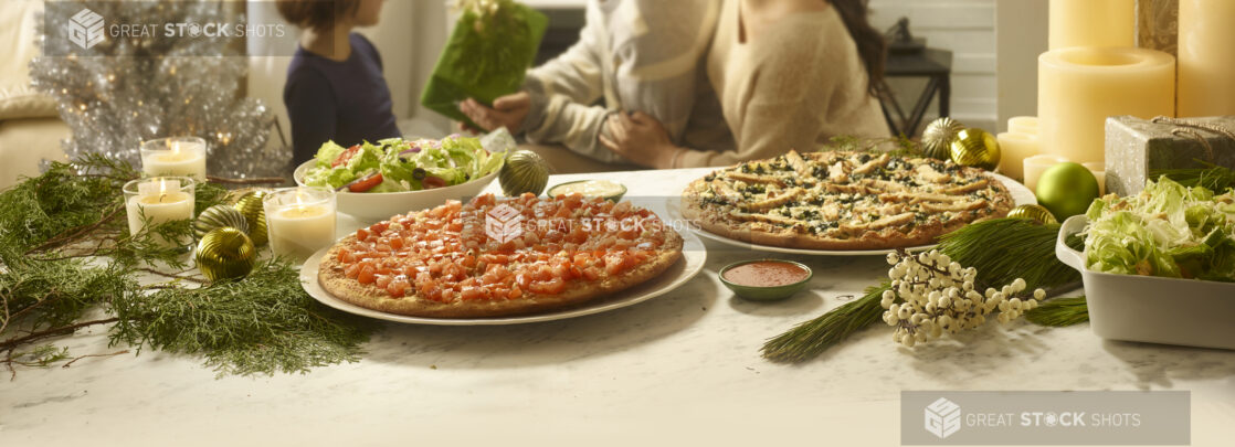 Two pizzas and a salad on a table surrounded by holiday decor with a family exchanging gifts in the background