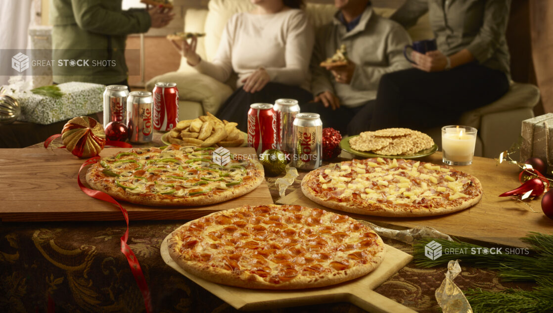 Various whole pizzas on wooden boards with a side of potato wedges, pizzelle and cans of pop with a family eating pizza in the background during the holidays