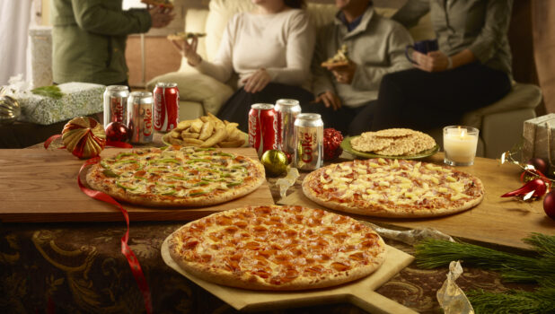 Various whole pizzas on wooden boards with a side of potato wedges, pizzelle and cans of pop with a family eating pizza in the background during the holidays