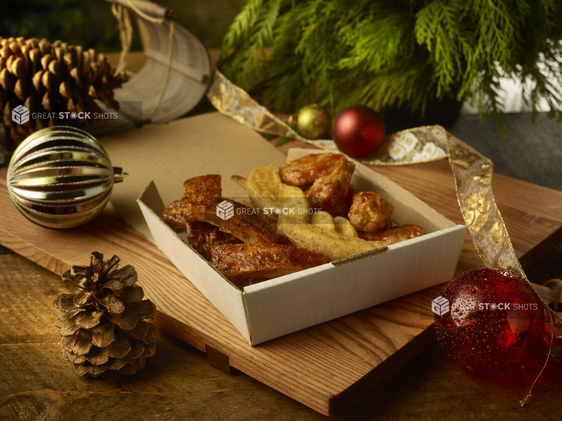 Order of ribs, potato wedges and chicken wings in a takeout container on a wooden board surrounded by festive decorations