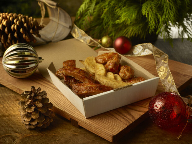Order of ribs, potato wedges and chicken wings in a takeout container on a wooden board surrounded by festive decorations