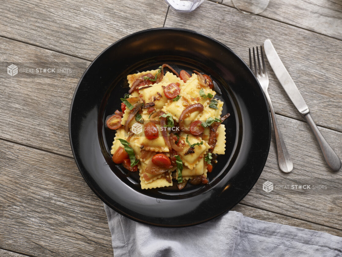 Square ravioli with grape tomatoes, red onion and fresh basil in a black pasta bowl on a grey wooden background