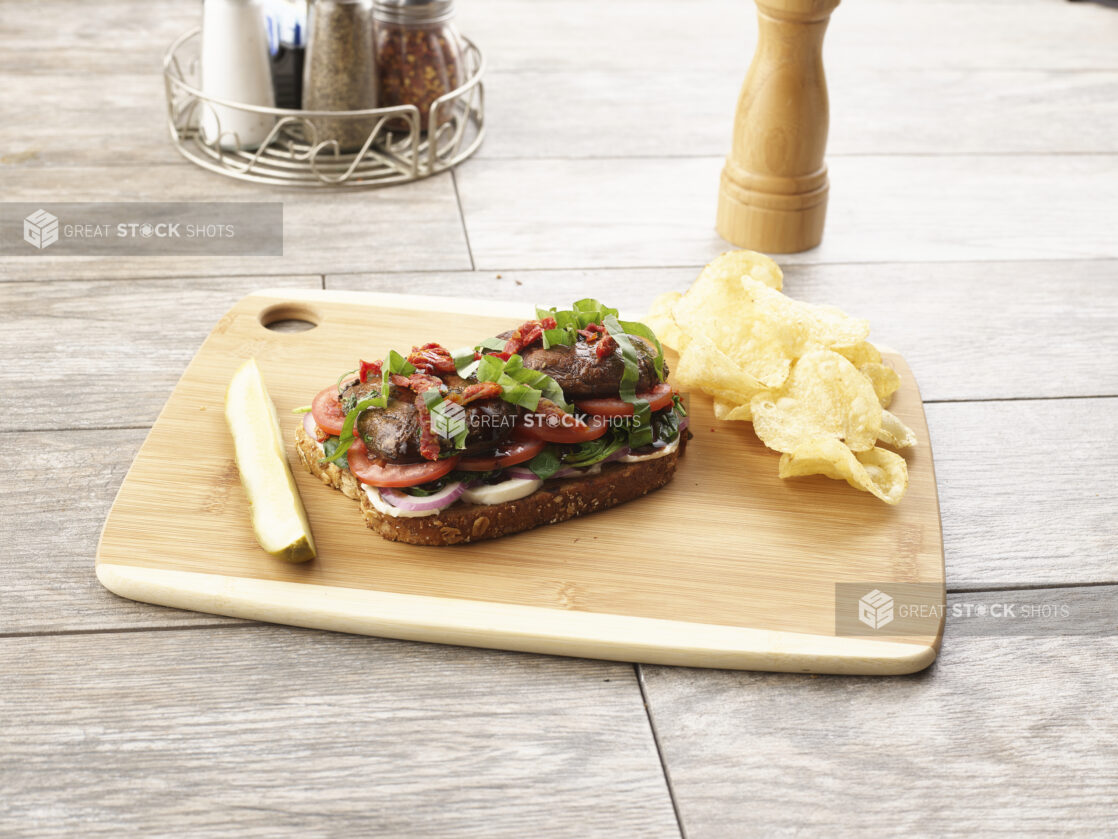 Open faced portobello mushroom sandwich with tomatoes, red onion and sundried tomatoes on multigrain bread on a wooden board with a pickle and potato chips on the side