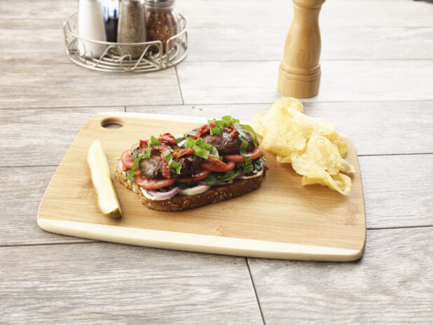 Open faced portobello mushroom sandwich with tomatoes, red onion and sundried tomatoes on multigrain bread on a wooden board with a pickle and potato chips on the side