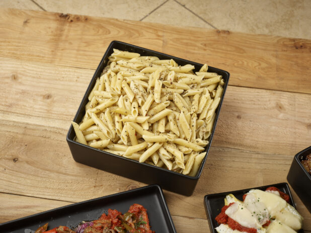 Penne pasta in a black square bowl with other Italian dishes in the foreground on a wooden background