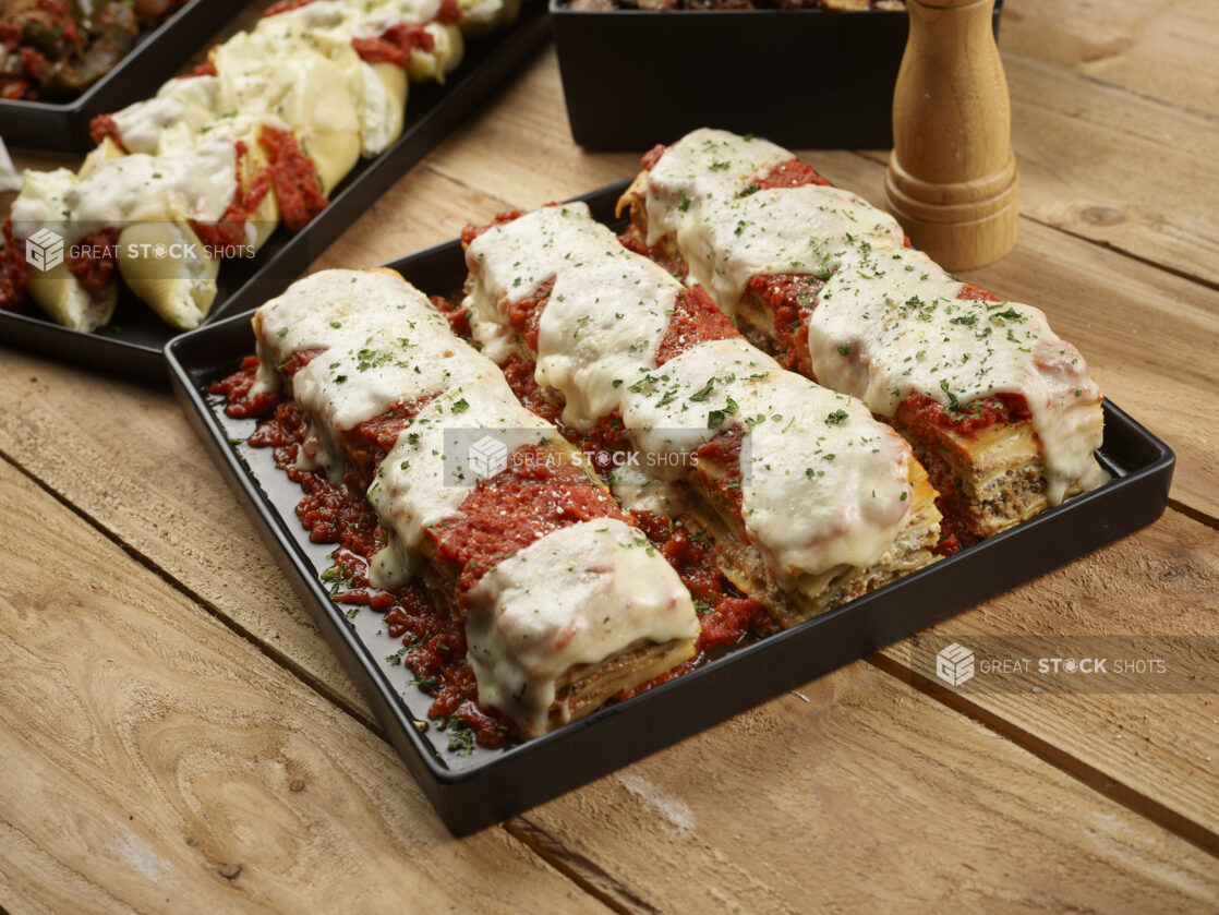 Lasagna on a black square platter with stuffed pasta shells on a rectangular black platter in the background on a wood tabletop